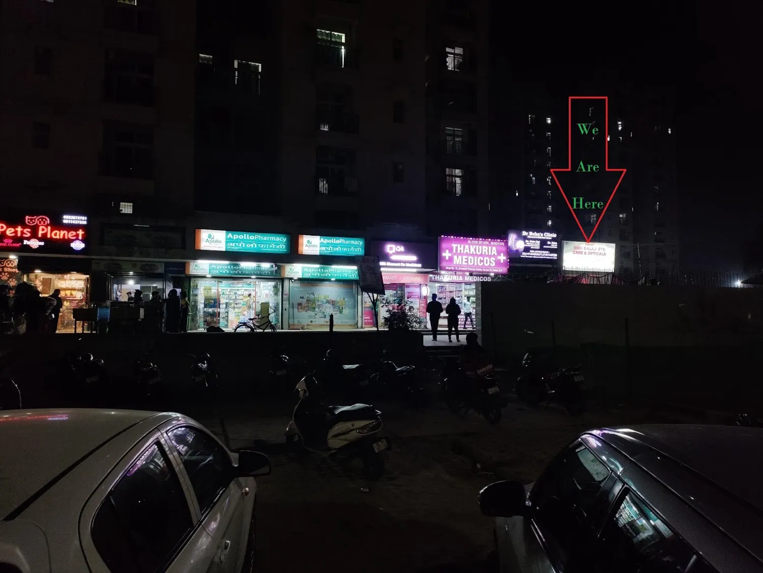 The shopping row at night. Shri Balaji Eye Care and Opticals is the lit sign at the right-hand end, past Apollo Pharmacy and Thakuria Medicos.