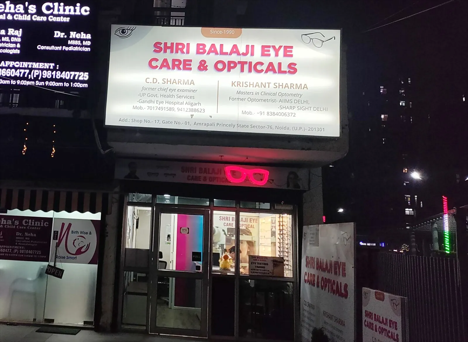 The illuminated sign over the door: Shri Balaji Eye Care and Opticals, since 1990, with both practitioners' names and where each of them trained.